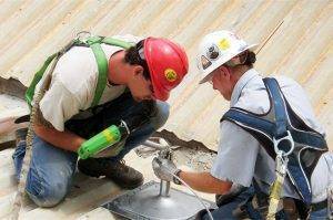 two commercial roofers wearing fall protection harnesses install a single-point anchor